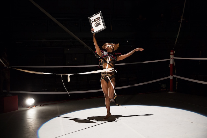 chipaumire holding a sign that says 'round one' in a boxing ring. Ropes encircle her stomach. 